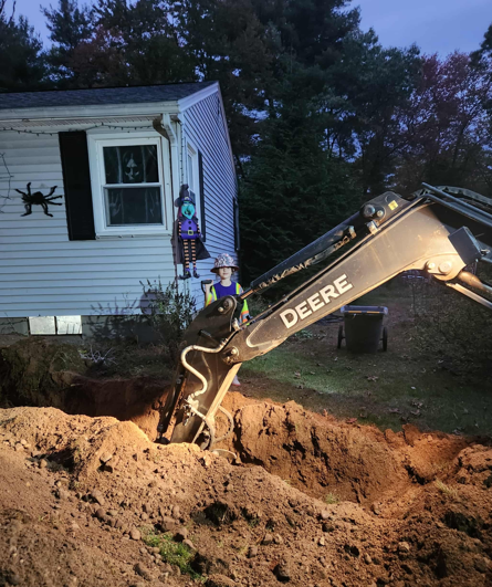 Excavator digging a trench beside a white house at dusk, with dirt piled in the foreground.
