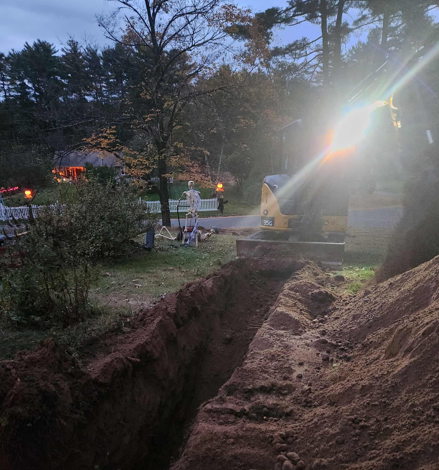 Freshly dug trench in a yard at dusk, with trees, a bench, and bright sunlight shining through.