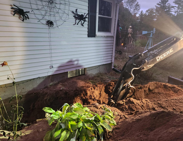 Excavator digging along a house foundation, with soil piled beside white siding and a window