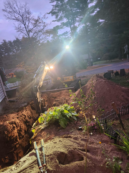 Construction trench at dusk with bright work lights, soil piles, and trees in the background