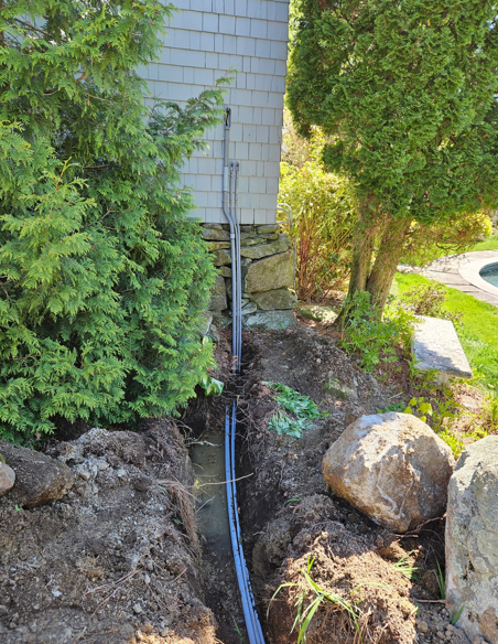 Narrow trench with a black pipe beside a house, bordered by dirt, rocks, and evergreen shrubs.