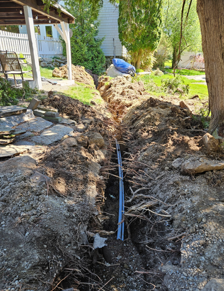 Trenched yard with exposed black cable running through muddy ground beside a house and trees