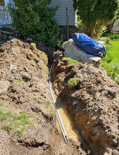 Trench dug beside a house with a pipe in the bottom and blue tarp-covered materials nearby