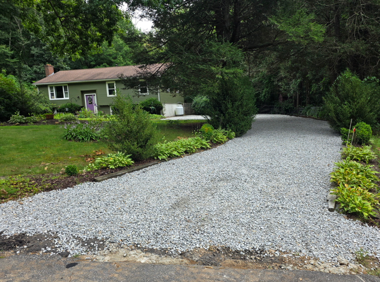 Gravel driveway curving to a small house amid lush green trees and shrubs