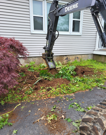 Excavator digging up a muddy yard beside a house with scattered uprooted plants.