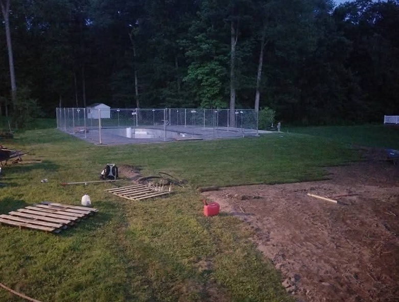 Chain-link animal pen at dusk beside a grassy yard, with scattered pallets and a red bucket in the foreground