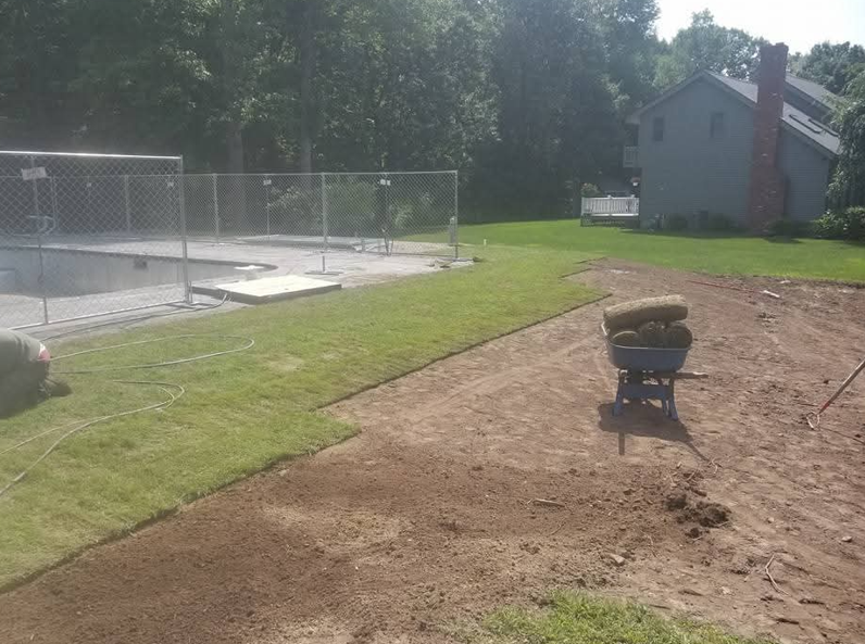 Backyard with fenced pool area, grassy lawn, and a wheelbarrow on a dirt patch near a house