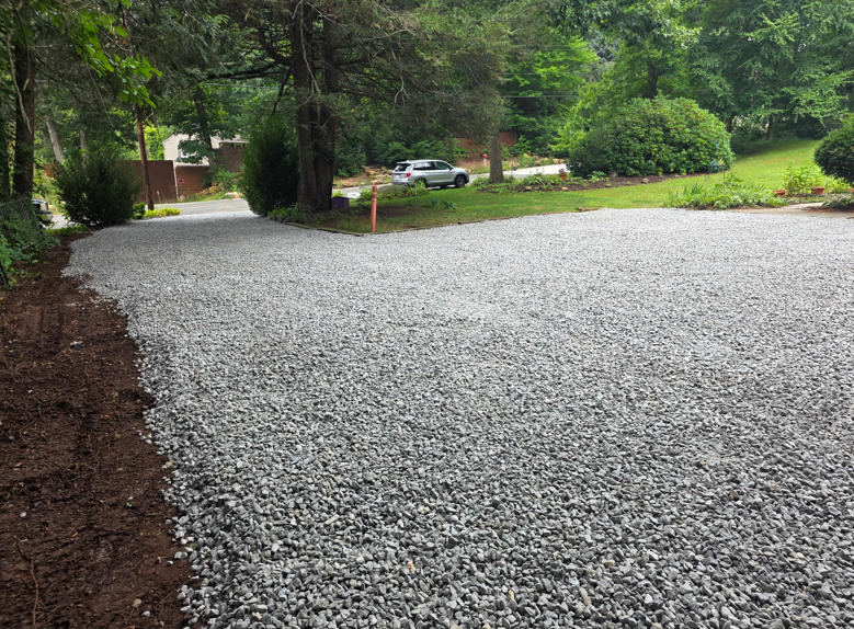 Gravel driveway under construction beside a wooded residential road on a rainy day