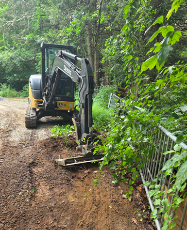 Mini excavator clearing muddy roadside beside a green forest fence.