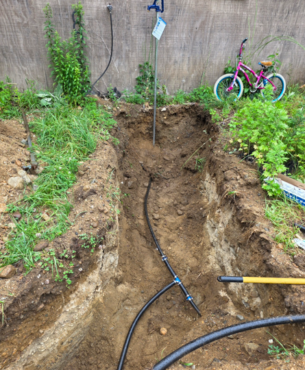 Dug trench in a backyard with black hose or cable running through it, near a wall and a pink bicycle.