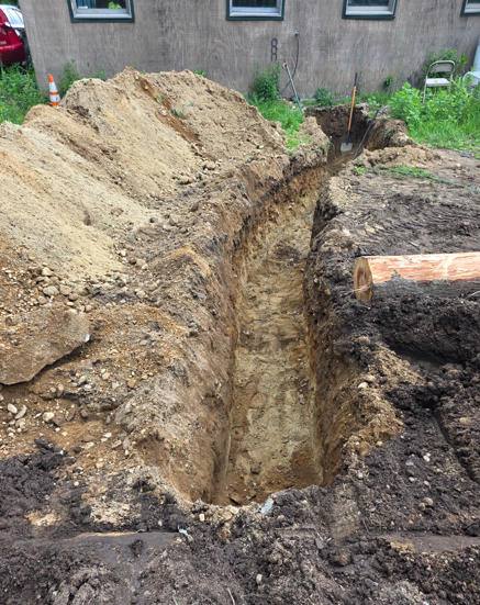 Deep trench dug through soil beside a building, with dirt piled along the edges and a wooden plank nearby.
