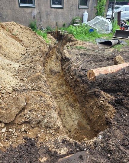 Narrow trench dug beside a house, with piles of soil and construction debris around it.