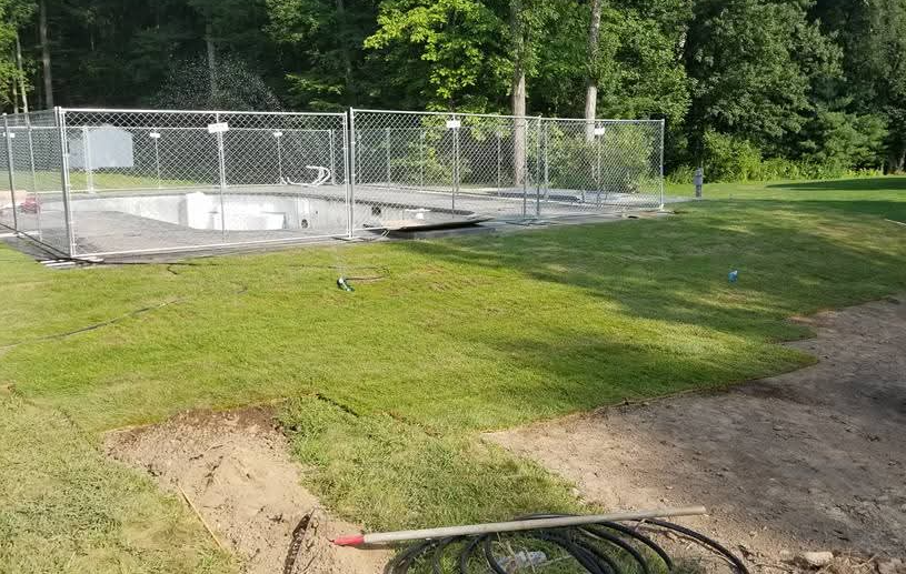 Grassy yard with a chain-link fenced utility area and dirt driveway in sunlight