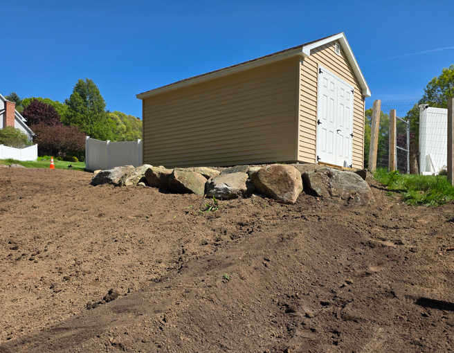 Beige shed on a dirt slope with rocks, under a clear blue sky.