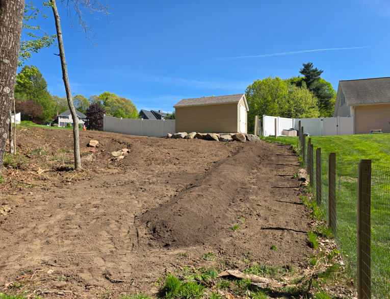 Bare dirt hillside beside a fence, with a small shed and trees under a clear blue sky.