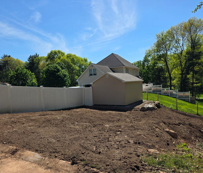 Freshly cleared backyard with dirt, white fence, beige shed, and house under a blue sky