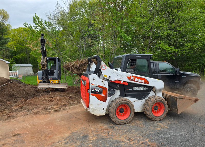 Bobcat skid steer lifting dirt beside a black pickup on a muddy driveway, with trees in the background