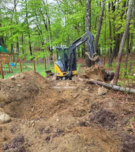 Mini excavator digging dirt in a wooded area, clearing around fallen logs.