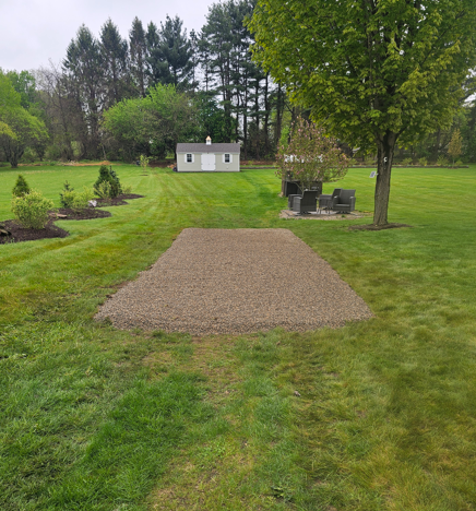 Backyard lawn with a gravel patio area, trees, and a small shed in the distance