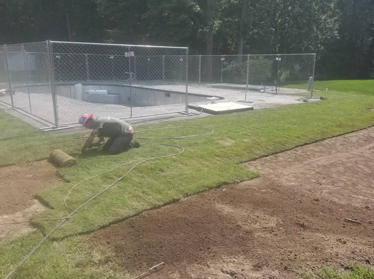 Worker on grassy slope using a hose near a fenced-in pool area.