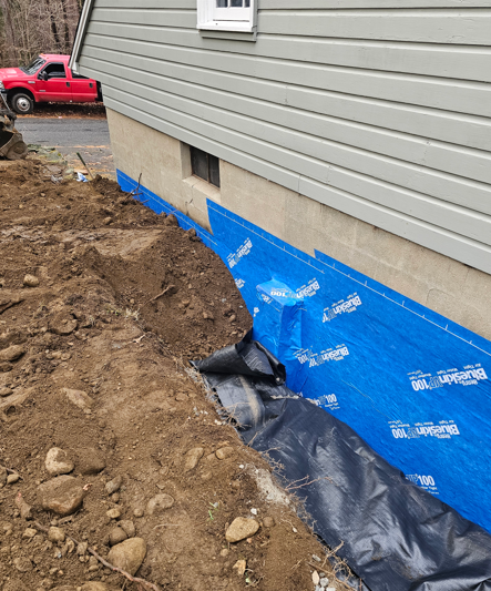 Blue house foundation wrapped in waterproofing, with dirt trench and black drainage fabric along the base.