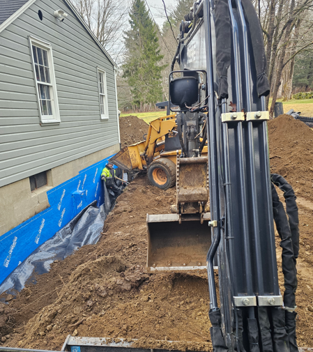 Excavator digging along a house foundation with blue tarp and piles of dirt in a yard