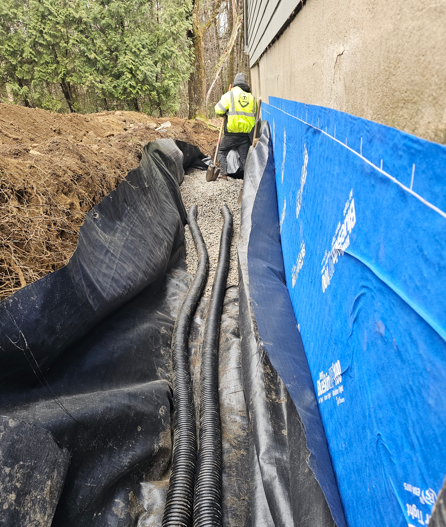 Worker on a narrow trench beside a building, with black drainage fabric and blue tarp lining the wall.