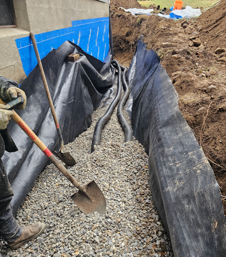 Construction trench lined with black plastic and blue boards, filled with gravel, with a shovel in the foreground