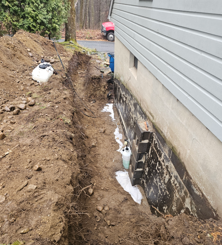 Trench dug along house foundation beside gray siding, with exposed black waterproofing and soil piles.