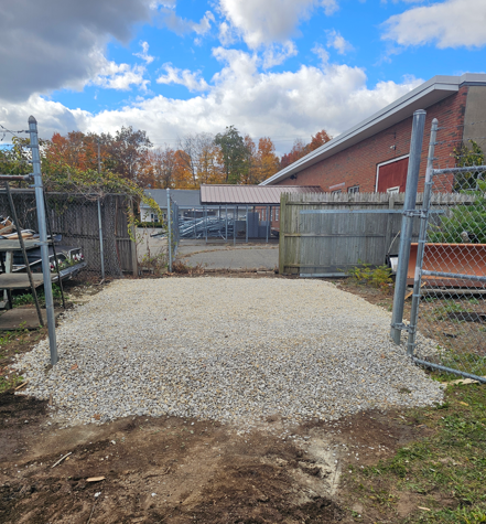 Gravel yard entrance with open chain-link gates, wooden fence, and brick building under a cloudy sky
