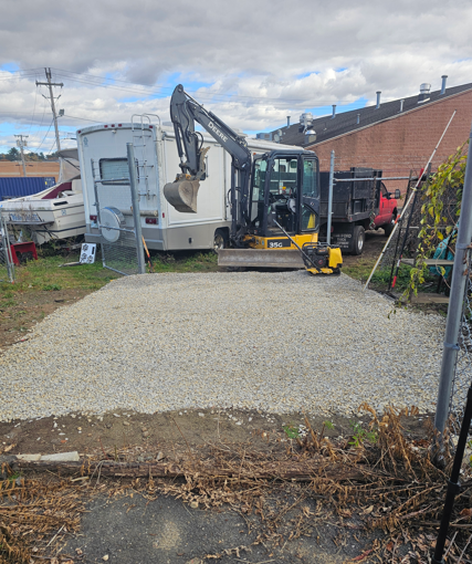 Mini excavator dumping gravel onto a driveway beside a house and trailer on a cloudy day