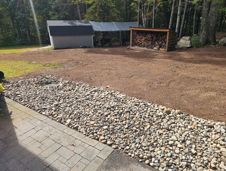 Backyard with gravel border, bare dirt area, shed, and wood storage under trees