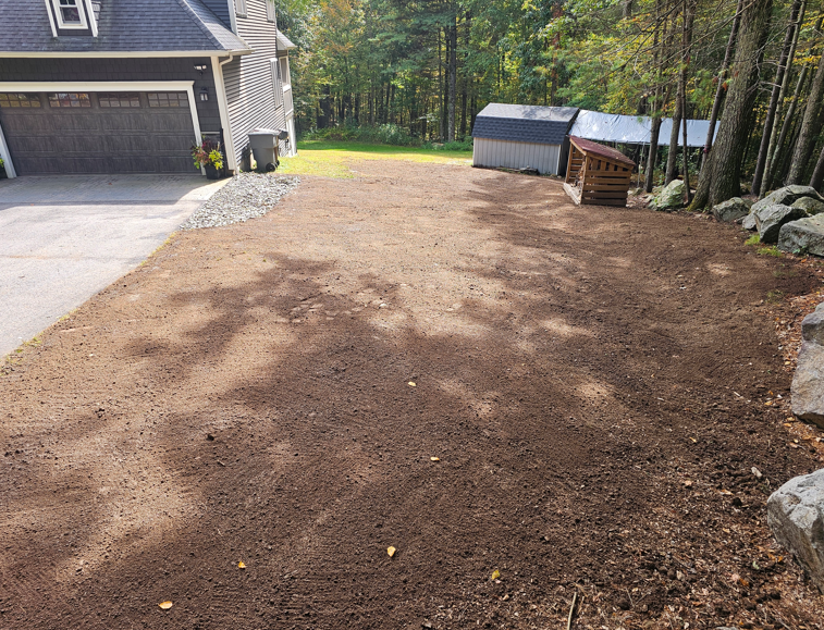 Freshly leveled dirt yard beside a house and driveway, with a shed and trees in the background