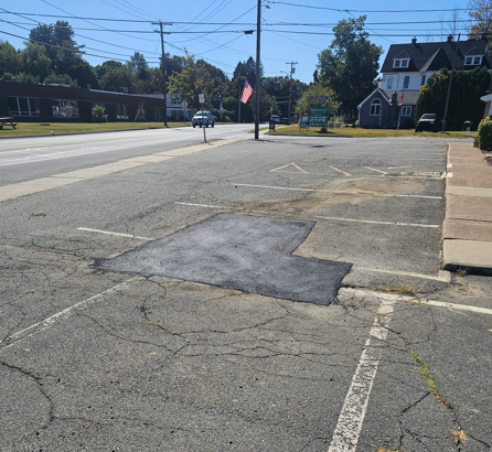 Cracked paved parking area beside a road, with patched asphalt and a building in the background.