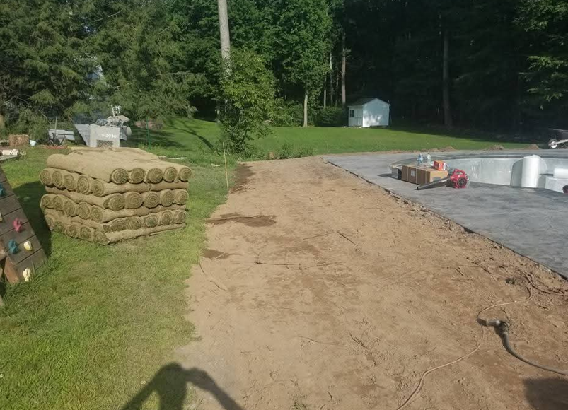 Dirt driveway beside stacked hay bales, paved area, and a small shed near trees.