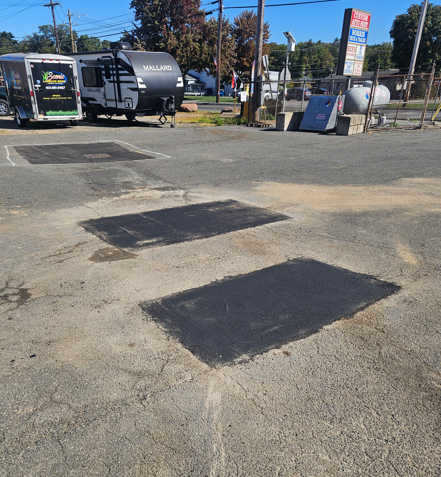 Cracked asphalt lot with dark pavement patches, parked trucks, and roadside signage on a sunny day