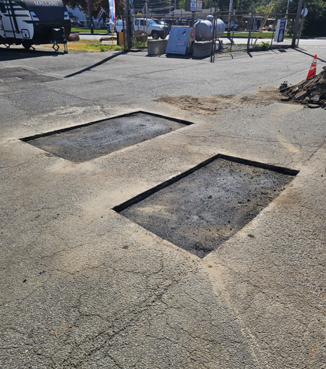 Two open rectangular pavement cutouts on a cracked asphalt lot near a road and sidewalk.