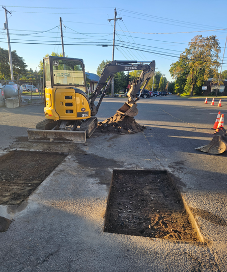 Yellow excavator patching a road trench at a street intersection with traffic cones nearby