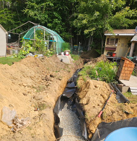A narrow trench with black lining cuts through a dirt yard beside houses and a green garden tunnel.