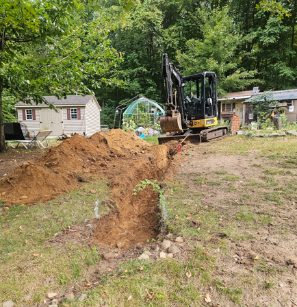 Backhoe digging a trench in a grassy yard beside a pile of dirt and a small house