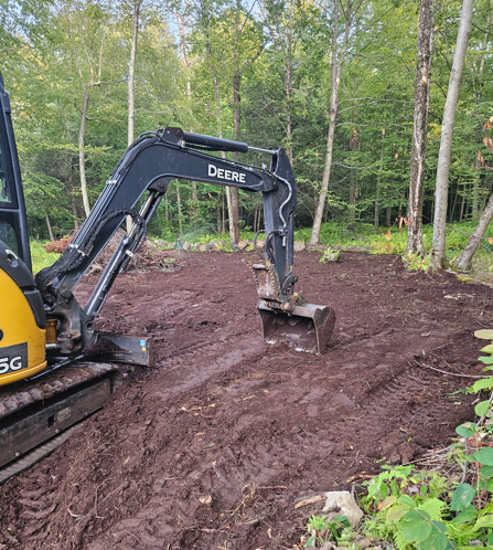 Deere excavator grading muddy ground beside a wooded area