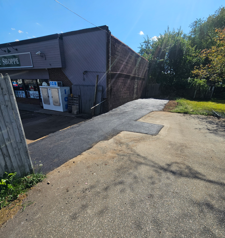 Sunlit paved path beside a brick building and fenced area, leading toward trees and grass.