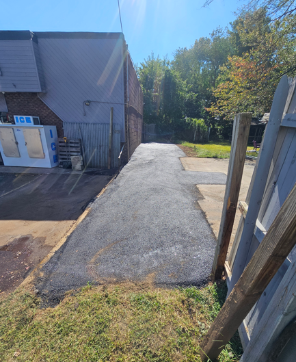 Paved driveway beside a brick building, leading to trees and a blue sky in a sunny outdoor area
