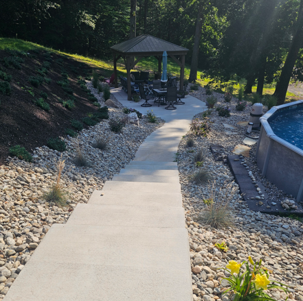Concrete path leading to a gazebo in a landscaped backyard with rocks, trees, and a pool edge