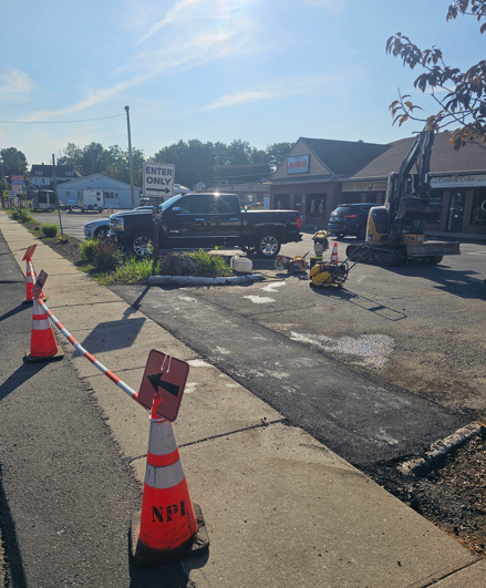Roadwork scene with orange cones and caution tape around a damaged curb, truck and excavator nearby.