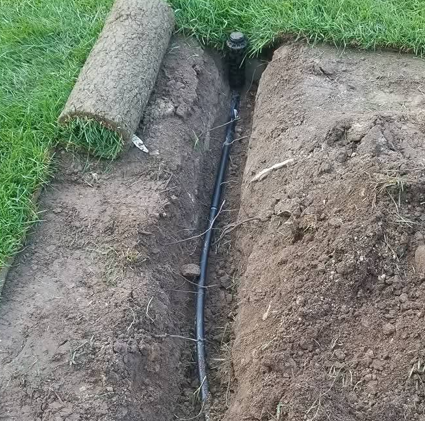 Freshly dug trench with black hose or cable laid between rolled turf and soil in a grassy yard