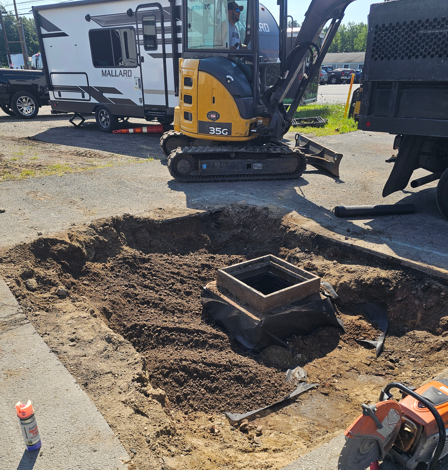 Excavator at a dug-up construction site around a square utility box, with tools and equipment nearby.
