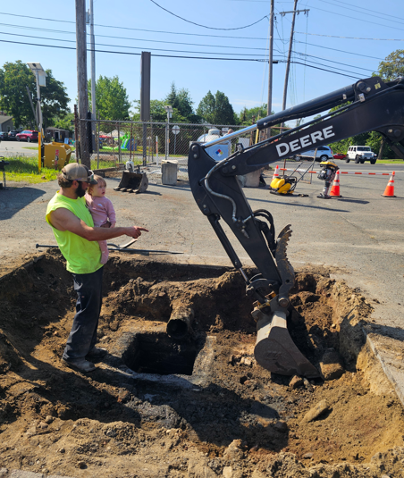 Worker directs excavator digging a trench beside a road with orange traffic cones nearby.