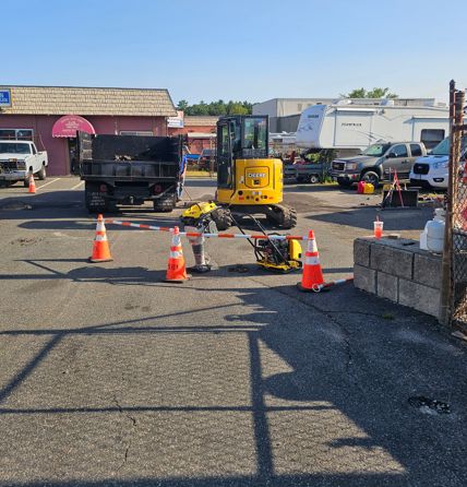 Parking lot roadwork with orange cones, a yellow compactor, and dump trucks near storefronts
