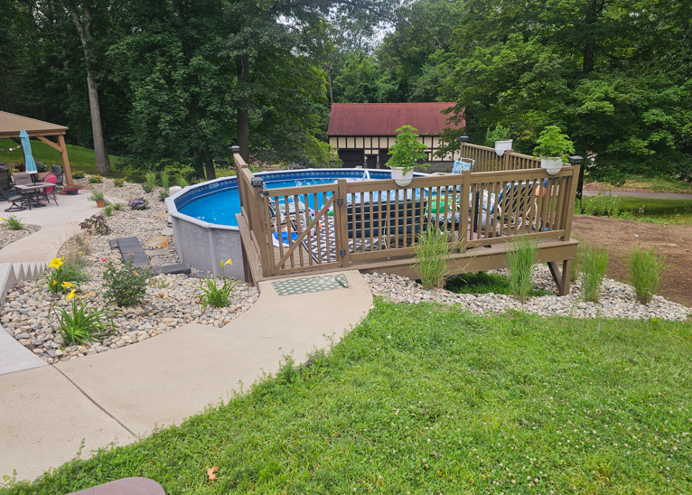Backyard above-ground pool with wooden deck, surrounded by garden beds and lawn